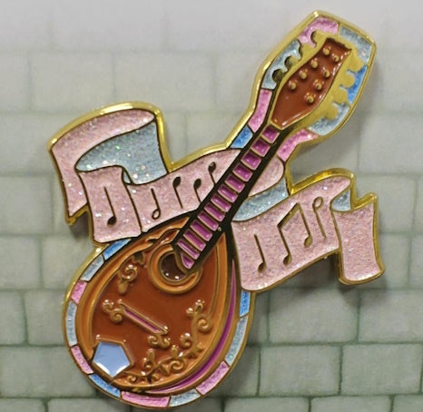 Soft enamel pin in the shape of a bard's mandolin, with scroll detail on the body and pink frets along the neck. It's surrounded by a thin stained glass background in the shades of pink, blue & white. There is also a pink scroll with musical notes flowing behind the neck of the mandolin. The stained glass area and music scroll have pink, blue and white glitter.